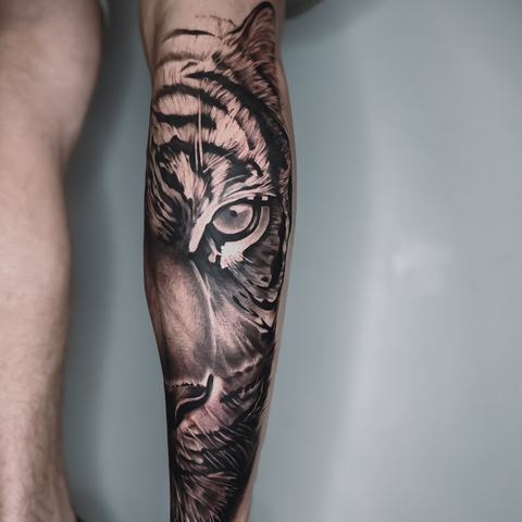 A realistic tiger portrait runs along the lower leg, using finely worked fur, sharp stripes and deep shadows to emphasize the animal’s intense expression.